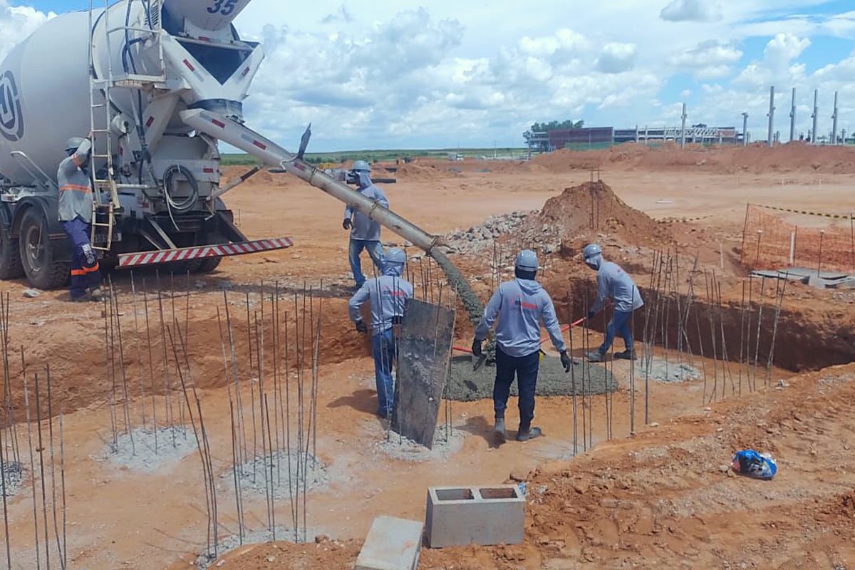 Equipe trabalha em fundações e caminhão betoneira ao fundo em canteiro de solo vermelho
