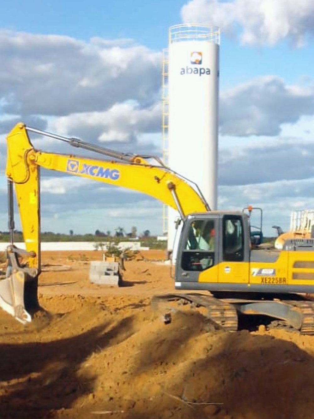 Escavadeira hidráulica em serviço de terraplenagem ao lado de silo do complexo