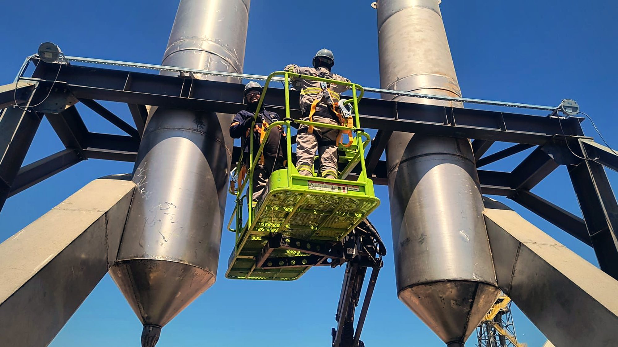 Dois eletricistas trabalham em plataforma suspensa entre dois silos metálicos, a vários metros do solo