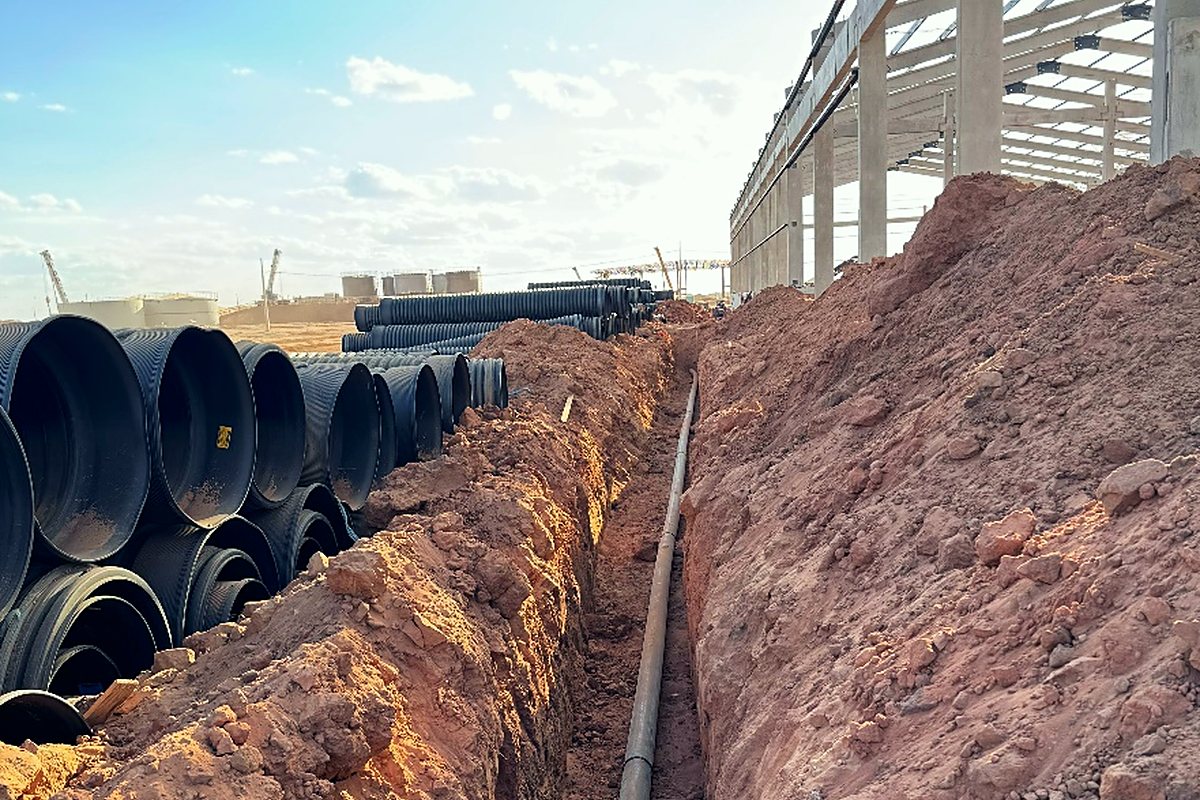 Dutos empilhados ao lado da vala, com estrutura metálica da planta industrial ao fundo